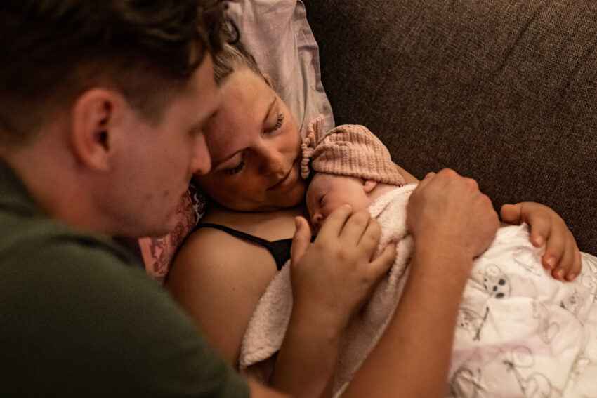 Na een mooie thuisbevalling ligt moeder met haar dochter op de bank en vader aait haar over haar wang, vastgelegd door geboortefotografie.
