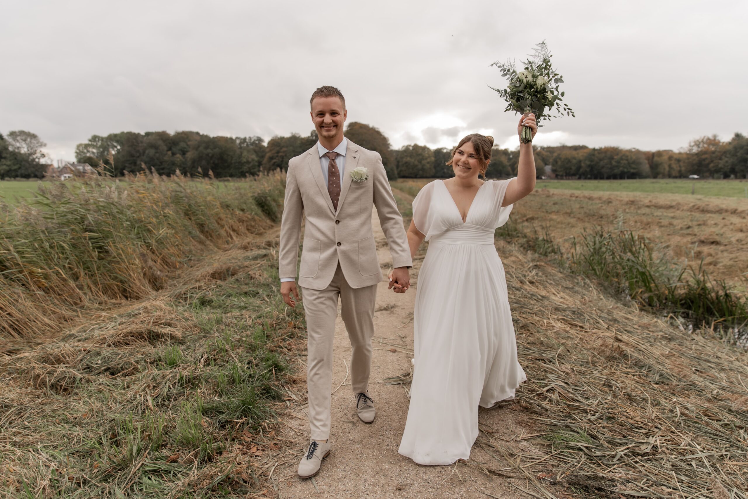 Trouwfotografie in Haarlem, bruidspaar loopt vrolijk door een weiland en de bruid heeft haar hand met boeket in de lucht.