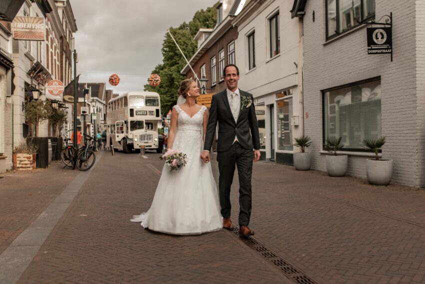 Trouwfotograaf in Zoetermeer, bruidspaar loopt door de straat naar de kerk, met op de achtergrond een engelse bus als trouwvervoer.
